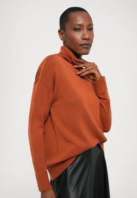 Oransje strikket genser med høyt ribbemasket krage, oversized passform, ribbestrikkede mansjetter og detalj i sidesømmen, kombinert med et svart skinnskjørt.