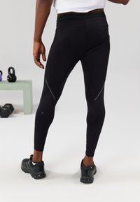 Juodos sportinės legginsai, turintys lygią tekstūrą, su atspindinčiais akcentais šonuose, derinami su juodais bėgimo batais.