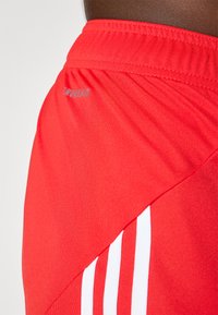 Röda sportshorts i mjukt tyg, med en elastisk midja, tre vita ränder på sidan och en liten logotyp i grått.
