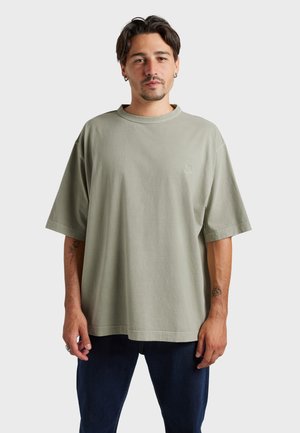 Homme aux cheveux courts et foncés et à la barbe, portant un t-shirt vert clair surdimensionné et un pantalon foncé, debout devant un fond uni clair.