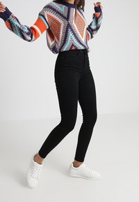 Suéter de punto multicolor con patrones geométricos en azul, naranja y blanco; combinado con jeans negros ajustados y zapatillas blancas.
