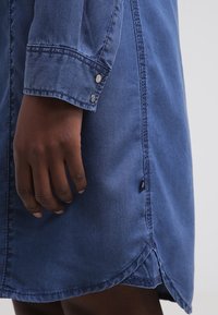 Abito in denim blu con polsini bottonati, orlo curvo e dettagli cuciti. Il tessuto appare morbido con una consistenza liscia.