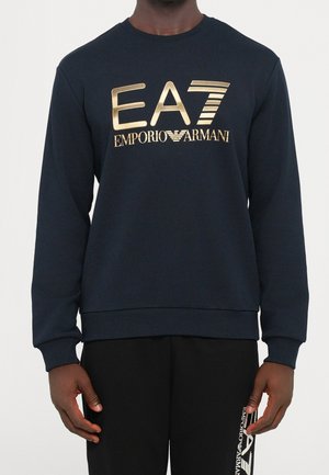 Personne portant un sweat-shirt bleu marine avec le logo doré "EA7 Emporio Armani" et un pantalon noir avec du texte de logo blanc sur le côté.