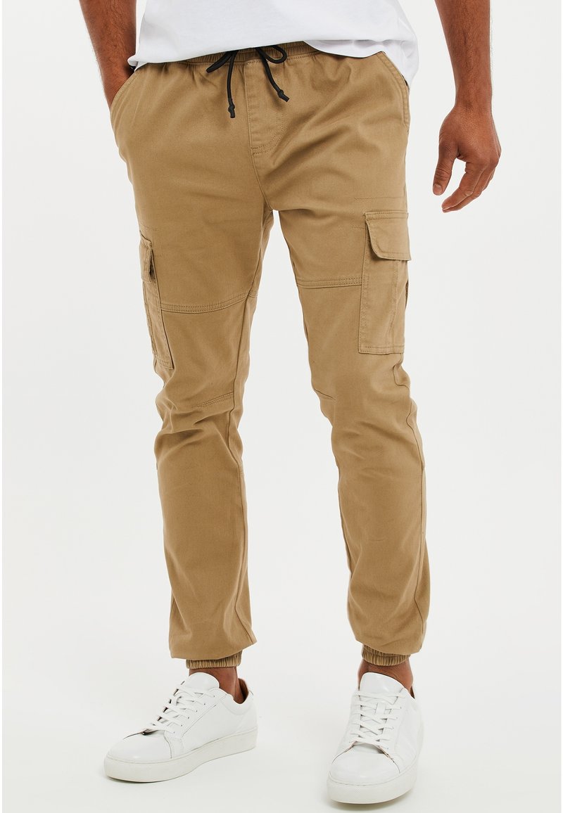 Homme portant un pantalon cargo jogger couleur beige avec taille à cordon et poches latérales, assorti à des baskets blanches et une chemise blanche.