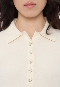 Cream-colored polo shirt with a soft texture, featuring a classic collar and six silver button accents along the front placket.