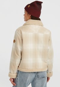 Person iført en beige ternet sherpa-jakke med pelskrave og manchetter, lyseblå jeans og en rød strikket hue, vendt væk.