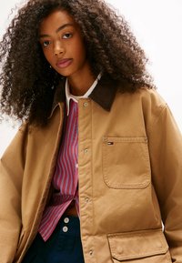 Brązowa kurtka z płótna o teksturowanym wykończeniu, z brązowym kołnierzem z sztruksu, kieszenią na piersi i małym logo. Pod kurtką koszula w paski.