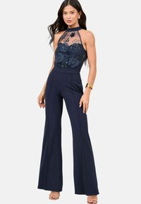 Navy jumpsuit with a floral lace bodice, halter neck, and wide-leg trousers. Features embroidered flowers and a textured detail.