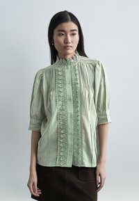 Γυναίκα που φοράει μπλούζα από ανοιχτό πράσινο δαντέλα με κουμπιά και φουσκωτά μανίκια, συνδυασμένη με σκουρόχρωμη φούστα, στέκεται μπροστά σε απλό φόντο.