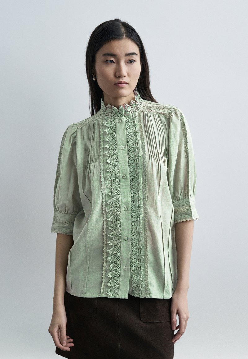 Γυναίκα που φοράει μπλούζα από ανοιχτό πράσινο δαντέλα με κουμπιά και φουσκωτά μανίκια, συνδυασμένη με σκουρόχρωμη φούστα, στέκεται μπροστά σε απλό φόντο.