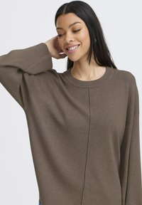 Pull en tricot marron avec un col rond, coupe décontractée, épaules tombantes et un motif de point texturé courant sur le devant.