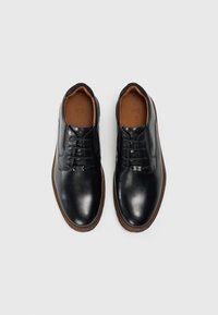 Un paio di scarpe eleganti in pelle nera con lacci, suola marrone e fodera interna color beige, viste dall'alto su uno sfondo bianco.