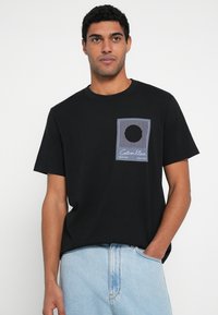 Melna kokvilnas T-krekls ar īsām piedurknēm un krūšu kabatu ar svītrotu grafisko dizainu un apli. Ikdienišķa piegriezuma un apaļa kakla izgriezuma krekls.