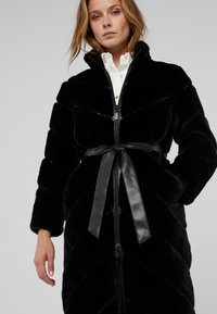 Cappotto in finta pelliccia nera con chiusura a zip, vita con cintura in pelle e motivo a zig-zag. Maniche lunghe e design con colletto alto.