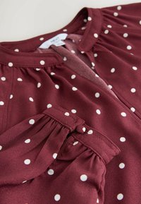 Blusa burdeos de tejido ligero con lunares blancos. Presenta un cuello redondo y detalles de mangas fruncidas aseguradas con un botón.