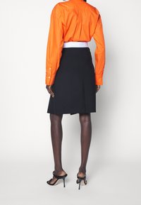 Musta hame, jossa on laskos takana, yhdistettynä oranssiin pitkähihaiseen nappipaitaan. Mustat remmilliset korkokengät täydentävät asun.