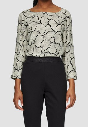 Donna che indossa una maglia a maniche lunghe con motivo astratto di petali in bianco e nero e pantaloni neri a vita alta su uno sfondo semplice.