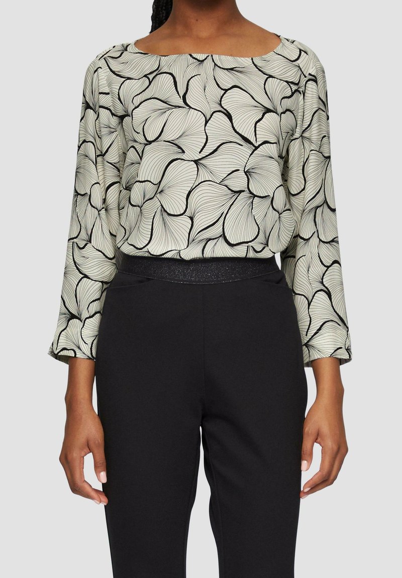 Femme portant un haut à manches longues avec un motif abstrait de pétales noir et blanc et un pantalon taille haute noir sur un fond uni.