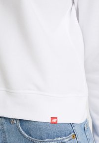 Gros plan d'une personne portant un sweat-shirt blanc avec une étiquette logo New Balance rouge au-dessus d'un jean en denim bleu.