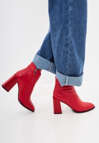 Bottes rouges à la cheville en cuir lisse, avec un talon épais et un détail de fermeture éclair. Associées à un pantalon en jean bleu retroussé.