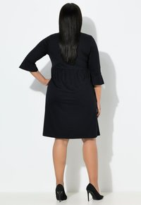 Žena s dlouhými černými vlasy, oblečená v černých šatech po kolena a černých vysokých podpatcích, stojící s jednou rukou na boku, otočená zády.