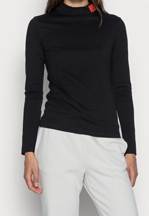 Žena nosí přiléhavý černý dlouhý rukáv s vysokým límcem a malou červenou visačkou u límce, v kombinaci s volnými bílými kalhotami.