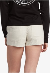 Beige cotton shorts with two back pockets, a clean finish, and a small heart accent. Worn with a black long-sleeve shirt.
