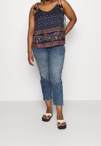 Camiseta de tirantes ajustables en un tono azul marino con estampado floral, que presenta borlas y un dobladillo estampado, combinada con unos jeans cortos de color azul claro y sandalias beige.