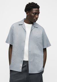 Νέος άνδρας που φοράει ένα γαλάζιο κοντομάνικο πουκάμισο με λευκή κεντητική πάνω από ένα λευκό μπλουζάκι και σκούρο παντελόνι, κοιτάζοντας προς τα δεξιά.