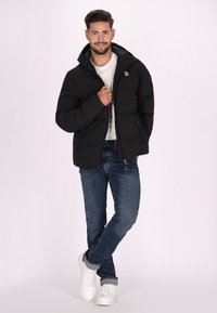 Giubbotto imbottito nero con cappuccio, dotato di chiusura con zip e due tasche laterali. Abbinato a jeans blu e sneakers bianche.