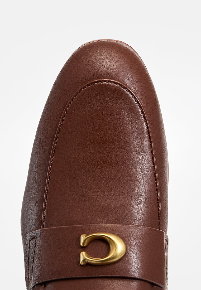 Mocassin en cuir marron avec un bout arrondi, présentant un accent matériel doré et une texture lisse, avec un minimum de surpiqûres sur le bord.