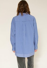 Chemise à manches longues avec des rayures verticales bleues et blanches, présentant une empiècement au dos, des poignets à boutons et une coupe décontractée. Tissu en coton lisse.