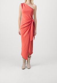 Femme portant une robe midi corail à une épaule avec un détail de nœud sur le côté, associée à des bottines en daim beige, se tenant devant un fond uni.