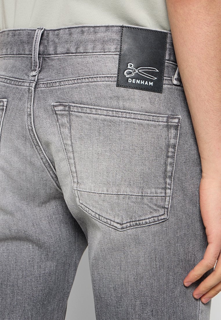 Grijze denim jeans met een leren label met het "DENHAM"-logo en een schaarafbeelding. Twee achterzakken zichtbaar en gestikte details.