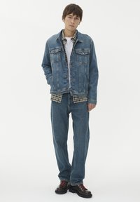 Mėlynas džinsinis švarkas su mygtukų užsegimu, dviem krūtinės kišenėmis, uždėtas virš raštuoto marškinio. Suderinta su plačiais mėlynais džinsais ir juodais batais.