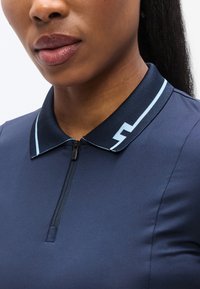 Πόλο μπλουζάκι σε ναυτικό μπλε με φερμουάρ στο γιακά, που διαθέτει αντίθετη ανοιχτό μπλε ρίγα και στυλιζαρισμένη λεπτομέρεια λογότυπου στον γιακά. Ομαλή υφή.
