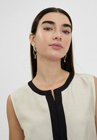 Blusa sin mangas en beige claro con un borde negro y detalle de botones. Los pendientes dorados presentan un diseño único y retorcido. Textura suave.