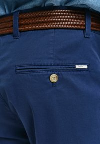 Navyblauwe katoen broek met een knoopvak op de achterkant, een gemerkte label en een bruine geweven leren riem. Zachte textuur met een subtiele glans.