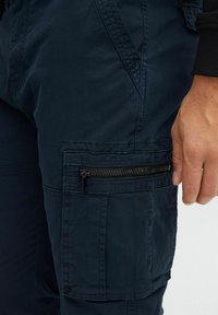 Granatowe spodnie cargo mają zasuwaną kieszeń na udzie, teksturowaną tkaninę oraz dopasowany pas, co podkreśla ich praktyczny design.