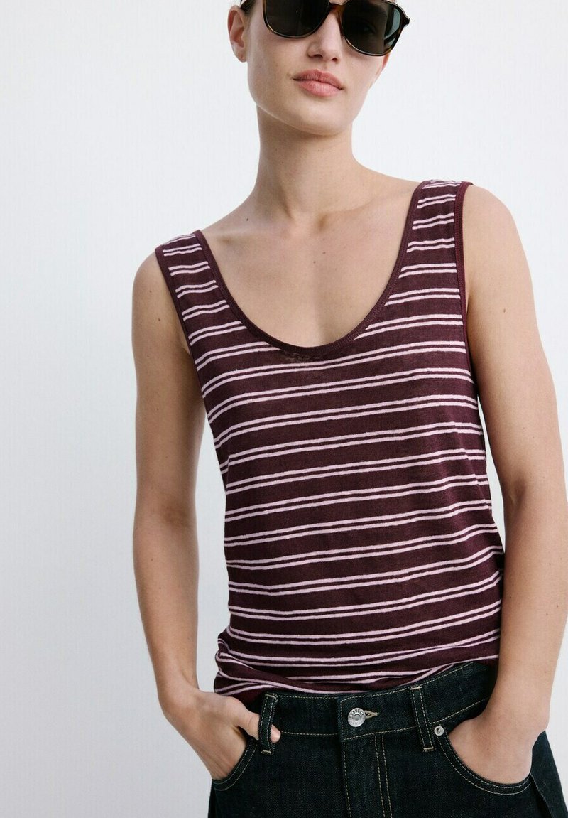 Femme portant des lunettes de soleil foncées, un haut sans manches rayé bordeaux et blanc, et un jean foncé, les mains dans les poches devant un fond uni.