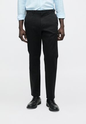 Férfi fekete szabott nadrágot, fekete bőr elegáns cipőt, és világoskék hosszú ujjú inget visel, melynek ujjai fel vannak hajtva.