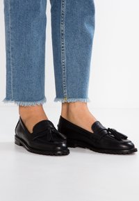 Zwarte leren instappers met kwastjes, gegolfde randen en een gestructureerde zool; gedragen met cropped blauwe denim jeans met rafelige zomen.