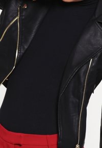 Chaqueta de moto de cuero negro con cremalleras doradas, combinada con un suéter de cuello alto azul marino ajustado y pantalones rojos, que exhibe texturas elegantes y fuertes contrastes.