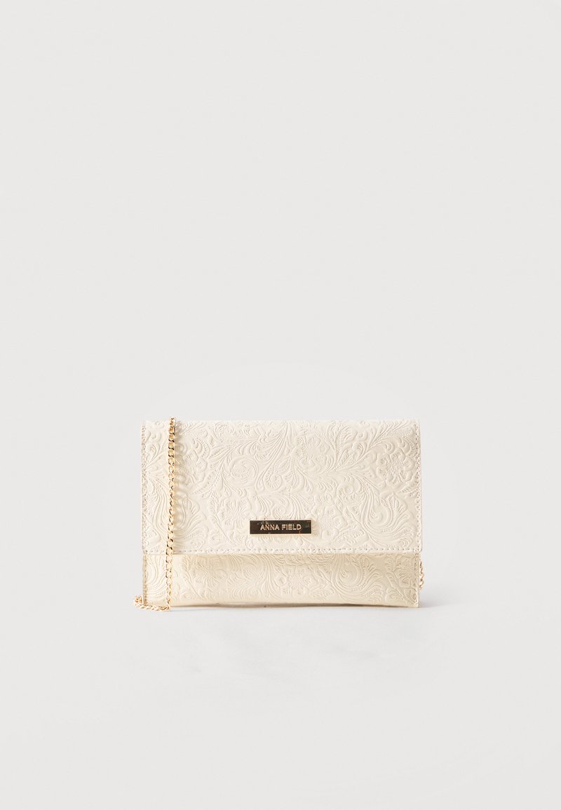 Pochette crème avec motif floral en relief, bandoulière chaîne dorée et petite plaque métallique portant l'inscription "ANNA FIELD" sur le rabat avant.