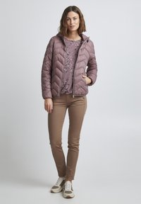 Veste matelassée violette clair avec une capuche, portée sur une chemise à motifs violette clair. Assortie à un pantalon ajusté marron et des baskets beiges.