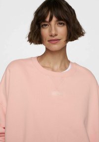 Sudadera rosa claro hecha de tela suave, con cuello de canalé, costuras sutiles y un pequeño logotipo cerca del escote.