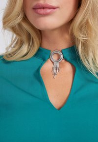Blusa teal con scollatura a occhiello, caratterizzata da una chiusura circolare in argento decorata con cristalli pendenti per un tocco in più. Tessuto dalla finitura liscia.