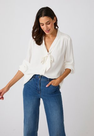 Frau trägt eine weiße Bluse mit langen Ärmeln und Frontband sowie blaue Jeans mit hoher Taille, steht vor einem schlichten hellen Hintergrund.