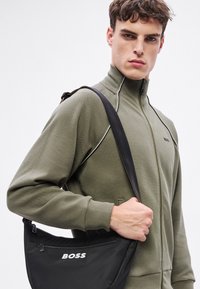 Χακί πράσινο μπουφάν με φερμουάρ και ραβδωτή υφή. Μαύρη τσάντα ώμου με λευκό λογότυπο "BOSS". Λείο συνθετικό υλικό, μινιμαλιστικός σχεδιασμός.