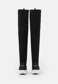 Botas negras hasta la rodilla hechas de tela elástica, con una superficie texturizada, suelas blancas acolchadas y un logo en el talón trasero.